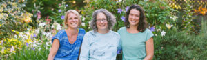 three smiling women in front of flowering bushes